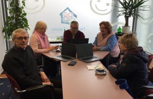 Ehrenamtliche und Besucherinnen und Besucher sitzen an einem Tisch darauf Computer